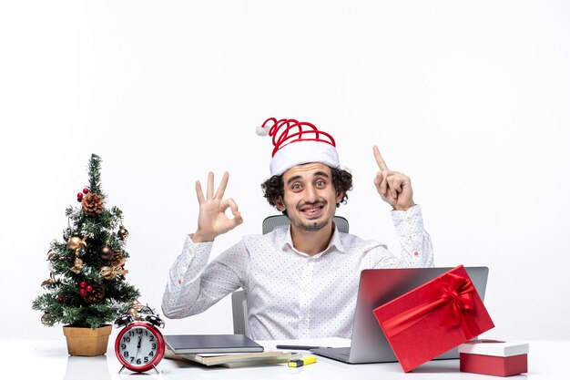 Счастливый позитивный молодой бизнесмен с забавной шляпой санта-клауса, указывая выше и делая жест очки в офисе на белом фоне