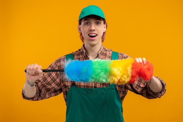 Happy and positive young cleaning man in plaid shirt jumpsuit and cap holding colorful duster looking smiling cheerfully