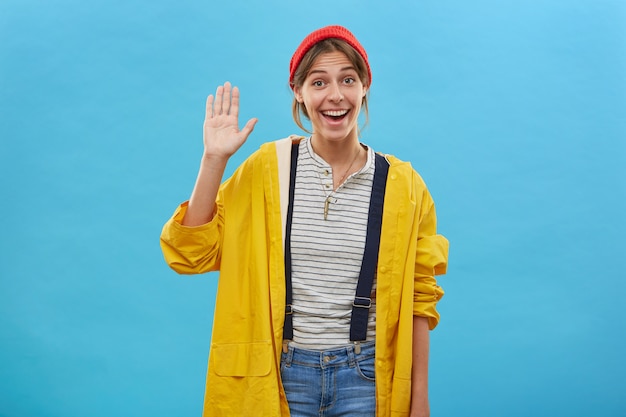 Free photo happy positive young female dressed casually waving with her palm greeting friends, showing friendly sign isolated over blue wall. smiling pleasant-looking woman raising her hand with pleasure