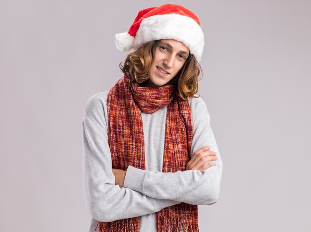 Happy and positive young man wearing christmas santa hat with warm scarf around his neck looking with smile on face with arms crossed 