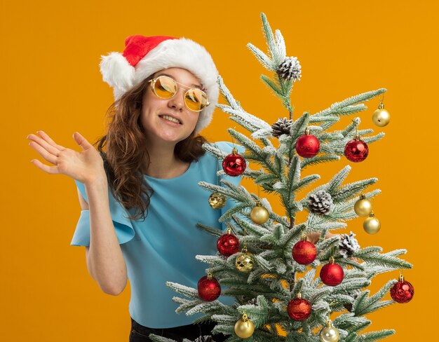 happy and positive young woman in blue top and santa hat wearing yellow glasses decorating christmas tree looking at camera waving with arm standing over orange background