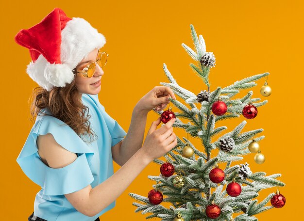 青いトップとオレンジ色の壁の上に立っているクリスマスツリーを飾る黄色いメガネを身に着けているサンタ帽子の幸せでポジティブな若い女性