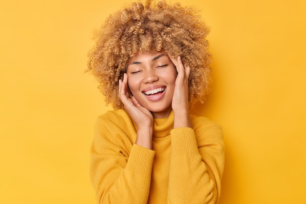 Felice giovane donna positiva con i capelli ricci chiude gli occhi si sente bene sorride ampiamente mostra denti bianchi vestiti con un maglione casual isolato su sfondo giallo vivido ricorda qualcosa di molto piacevole