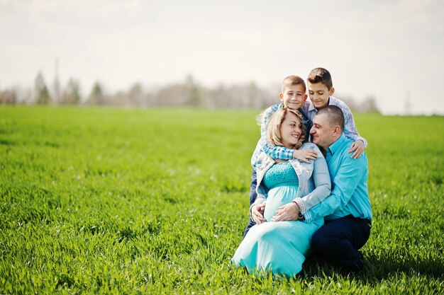 Счастливая беременная семья с двумя сыновьями, одетыми в бирюзовую одежду, проводят время вместе на зеленой траве в поле