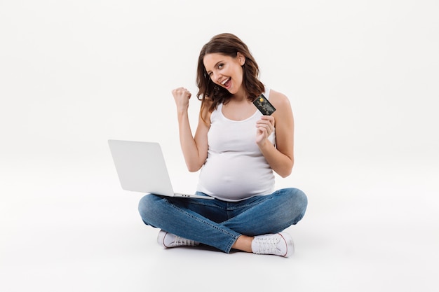 Happy pregnant woman holding debit card winking using laptop
