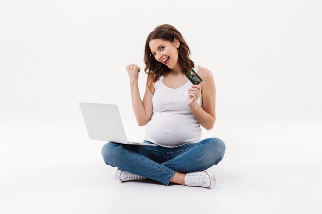 Happy pregnant woman holding debit card winking using laptop