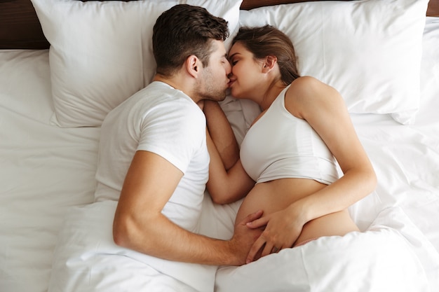 Happy pregnant woman lies in bed with her husband kissing.