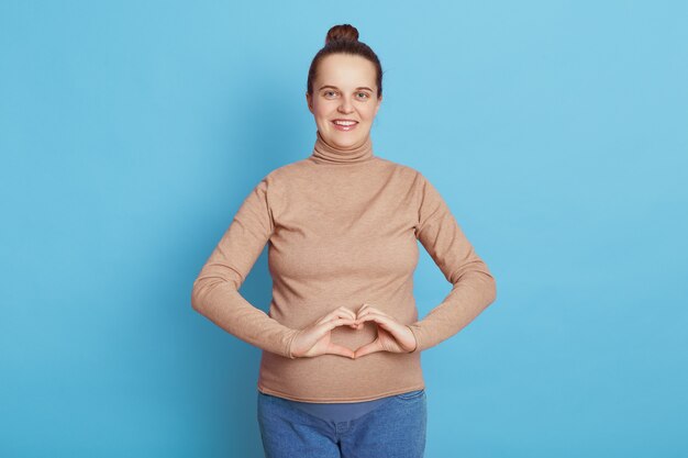 Happy pregnant woman making heart gesture in front of her belly, wearing casual attire, having hair bun, expecting mother standing isolated over blue wall.