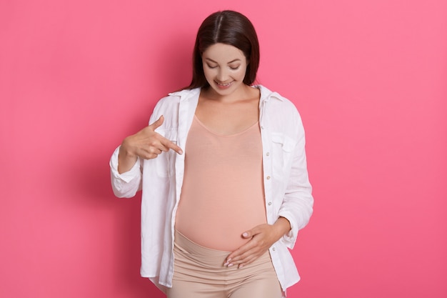 Happy pregnant woman pointing at her belly with smile and positive facial expression, wearing white shirt while standing against pink wall, dark haired expected mother.