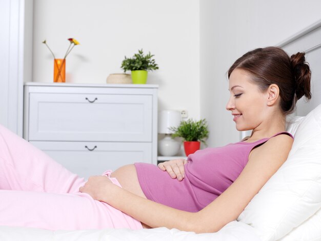 Happy pregnant young woman lying on the bed and touching belly