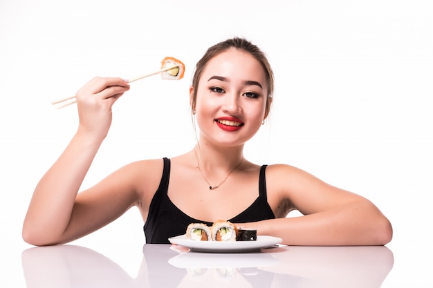 控えめな髪型でテーブルの上に座って幸せなかなりアジアの外観は、白で隔離される笑顔の巻き寿司を食べる
