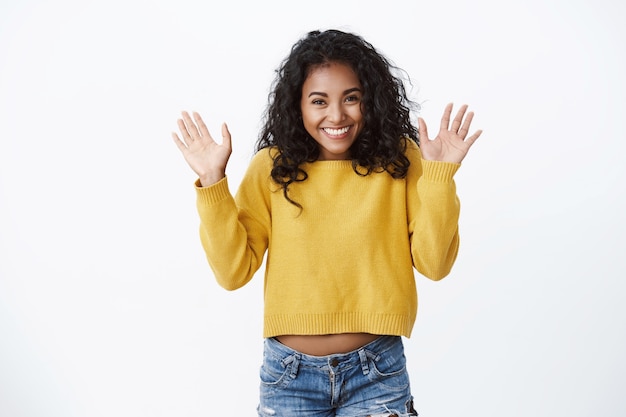 Happy pretty girl with curly hair showing excitement and happiness, greeting friends, waving raised palms hello, hi gesture, finally can wear new yellow sweater autumn chilly day