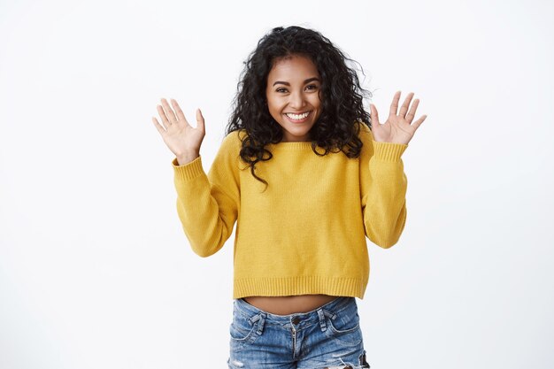 Happy pretty girl with curly hair showing excitement and happiness, greeting friends, waving raised palms hello, hi gesture, finally can wear new yellow sweater autumn chilly day