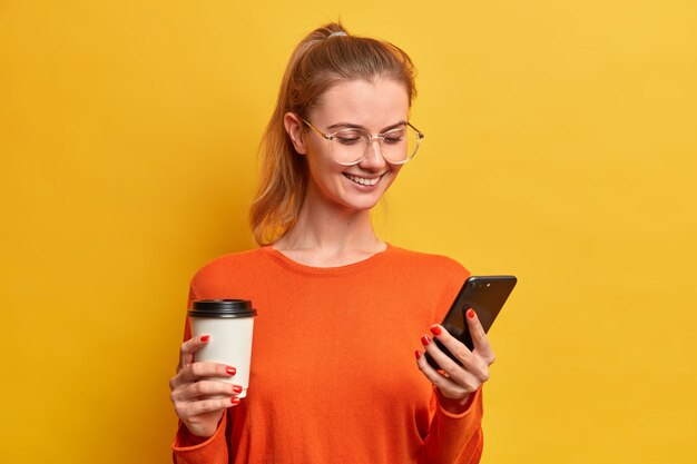 Happy pretty millennial girl downloads new mobile application, drinks coffee from paper cup, has pleasant smile, texting in chat, wears optical glasses, has combed hair in pony tail, surfs internet