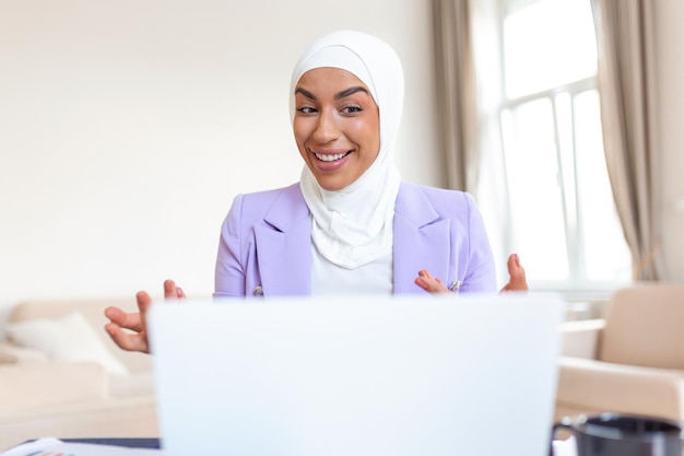 아늑한 소파에 앉아 노트북을 사용하는 행복한 예쁜 이슬람 아랍 여성 아름다운 젊은 이슬람 여성이 노트북을 사용하고 웃고 있습니다