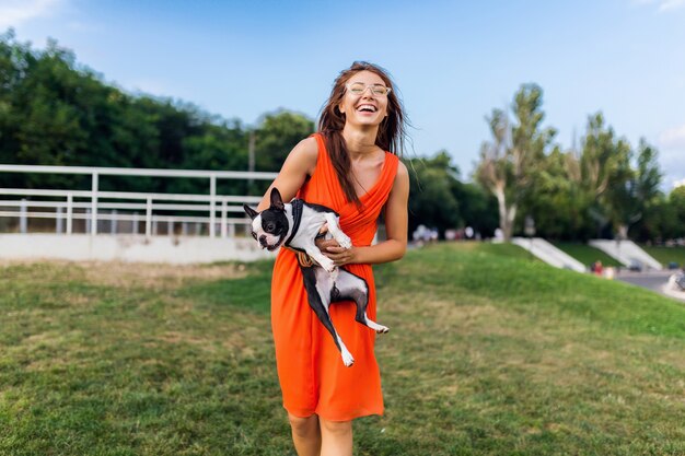 ボストンテリア犬を保持している幸せなきれいな女性公園、前向きなムードの笑顔、トレンディな夏のスタイル、オレンジ色のドレス、サングラス、ペットと遊ぶ、楽しんで、カラフル