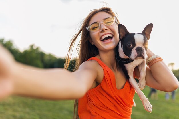 自撮り写真を作る幸せなきれいな女性公園、ボストンテリア犬を抱いて、ポジティブな気分を笑顔、トレンディな夏のスタイル、オレンジ色のドレス、サングラスを身に着けて、ペットと遊んで、楽しんでください