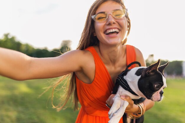 Счастливая симпатичная женщина в парке делает селфи, держит собаку бостон-терьера, улыбается позитивное настроение, модный летний стиль, носит оранжевое платье, солнцезащитные очки, играет с домашним животным, развлекается