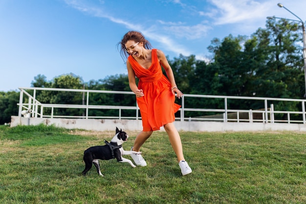 Happy pretty woman in park running with boston terrier dog, smiling positive mood, trendy summer style, wearing orange dress, playing with pet, having fun, colorful, active weekend vacation, sneakers