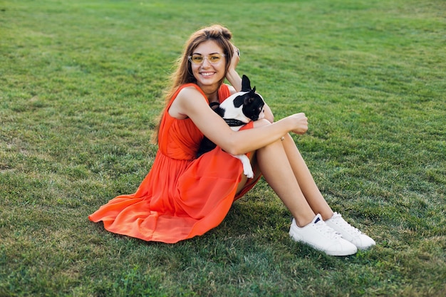Happy pretty woman sitting on grass in park, holding boston terrier dog, smiling positive mood, wearing orange dress, trendy style, slim legs, sneakers, playing with pet, summer fashion trend