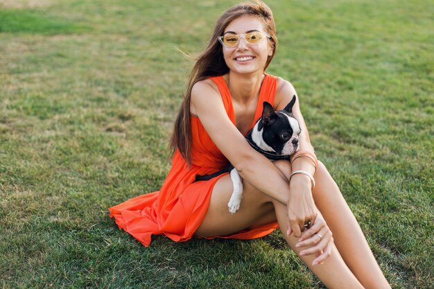 サマーパークの芝生に座って、ボストンテリア犬を抱いて、前向きな気分を笑顔、オレンジ色のドレスを着て、流行のスタイル、ペットと遊ぶ幸せなきれいな女性