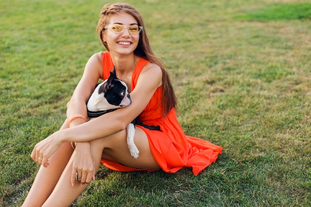 Счастливая красивая женщина, сидящая на траве в летнем парке, держа в руках собаку бостон-терьера, улыбаясь позитивным настроением, в оранжевом платье, модном стиле, играя с домашним животным
