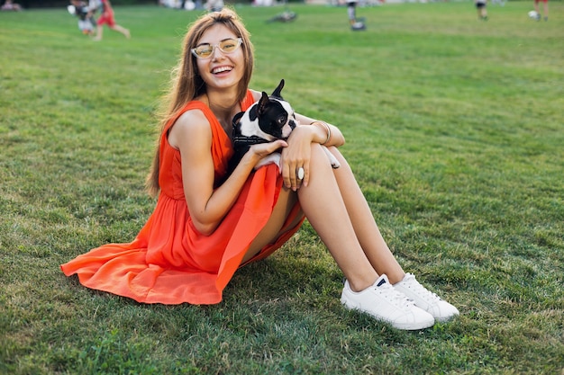 Счастливая симпатичная женщина, сидящая на траве в летнем парке, держа собаку бостон-терьера, улыбаясь позитивным настроением, в оранжевом платье, в модном стиле, стройные ноги, кроссовки, играя с домашним животным
