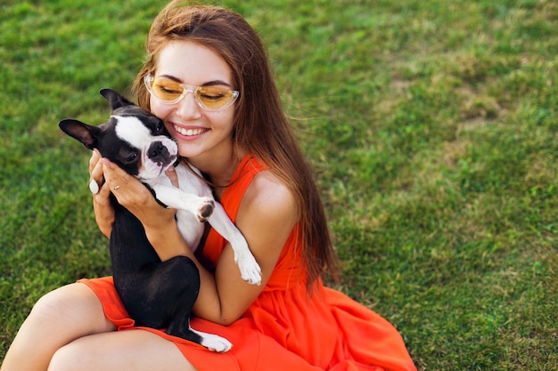 夏​の​公園​の​芝生​に​座って​、​ボストン​テリア​犬​を​抱いて​、​キス​、​オレンジ色​の​ドレス​を​着て​、​流行​の​スタイル​、​ペット​と​遊ぶ​幸せな​きれいな​女性