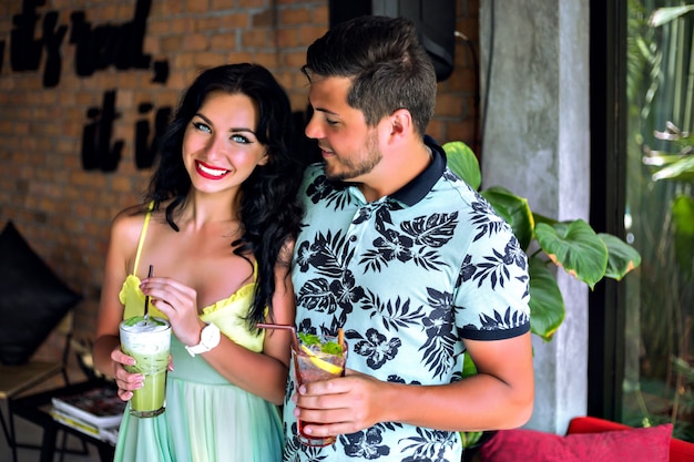 Happy pretty young couple enjoying their tasty sweet cocktail at tropical bar, color matching trendy clothes, summer vacation mood. Perfect romantic date.