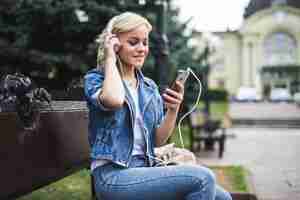 Free photo happy pretty young woman listening music in headphones and using smartphone while sitting on the bench in the city