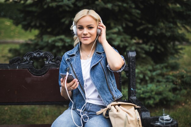 도시의 벤치에 앉아있는 동안 헤드폰에서 음악을 듣고 스마트 폰을 사용하는 행복 꽤 젊은 여자