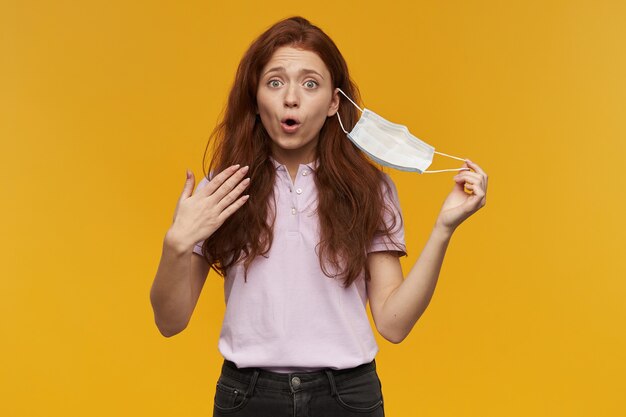 Happy pretty young woman taking off medical protective mask and taking a breathe of fresh air over yellow wall