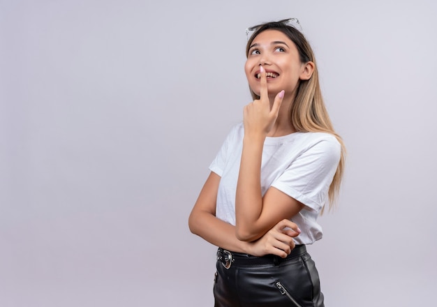 A happy pretty young woman in white t-shirt wearing sunglasses on her head keeping finger on her lips while thinking on a white wall