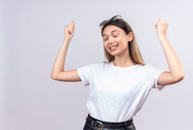 A happy pretty young woman in white t-shirt wearing sunglasses on her head raising clenched fists arm with closed eyes on a white wall