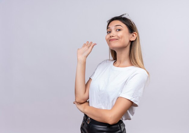 A happy pretty young woman in white t-shirt wearing sunglasses on her head raising her hand in goodbye gesture on a white wall