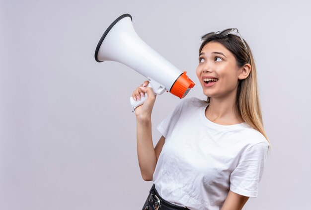 A happy pretty young woman in white t-shirt wearing sunglasses on her head speaking through megaphone on a white wall