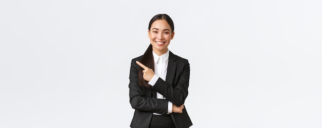 Happy professional asian female manager businesswoman in suit showing announcement smiling and pointing finger left at product or project banner standing white background