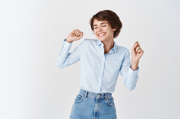 Happy professional woman feeling joy, dancing with closed eyes and cheerful smile, standing on white wall