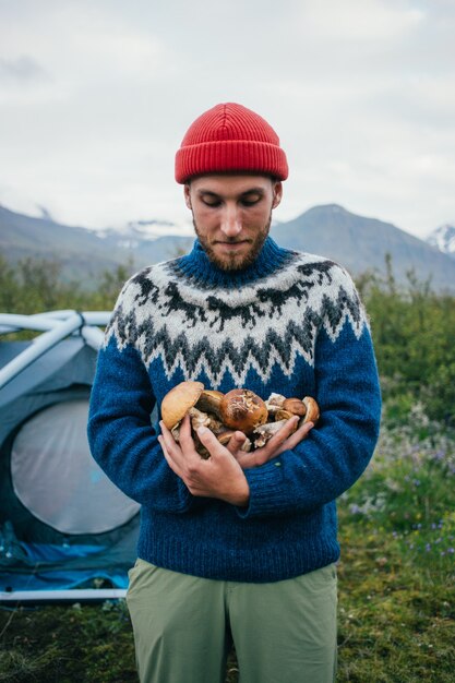 Happy, proud picker man in traditional blue wool sweater with ornaments stands on camping ground in mountains, holds in arms pile of delicious and organic mushrooms