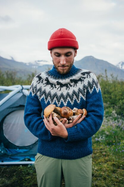 Счастливый, гордый сборщик в традиционном синем шерстяном свитере с орнаментом стоит на территории кемпинга в горах, держит в руках кучу вкусных и органических грибов