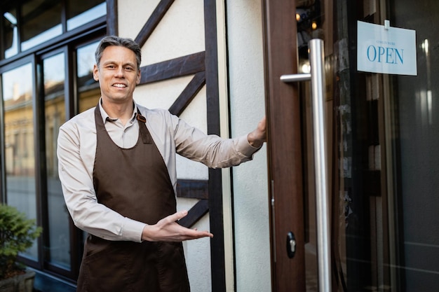 Happy pub owner welcoming guest while standing at entrance door and looking at camera.