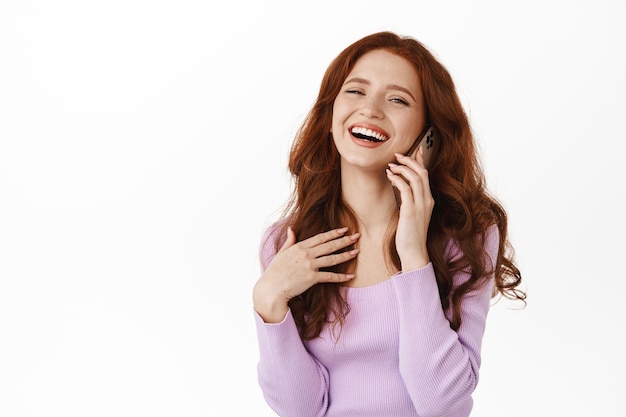 Happy redhead beauty girl talking on smartphone, laughing and smiling sincere while calling friend, communicating on mobile phone, standing against white background