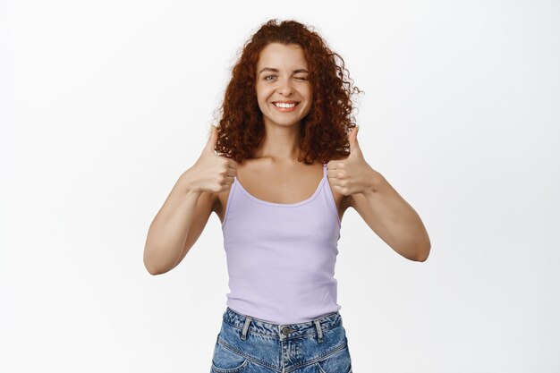 Happy redhead curly woman, showing thumbs up and smiling satisfied, recommending, leave positive feedback, like and agree on white