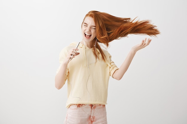 Free photo happy redhead girl playing karaoke app game on smartphone, singing song in earphones