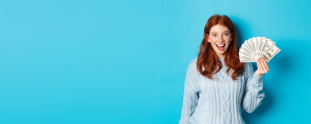 Free photo happy redhead girl in sweater staring excited at camera showing money dollars standing against blue