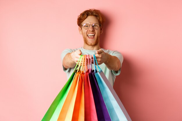 Happy redhead man stretch out hands with shopping bags, give you gifts, standing over pink background.