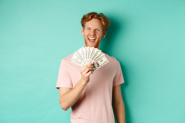 Happy redhead man in t-shirt showing money in dollars and smiling, making smug faces after winning cash, standing over turquoise background