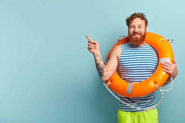 Happy redhead man with curly hair, relaxes at summer beach, poses with bright orange lifebuoy