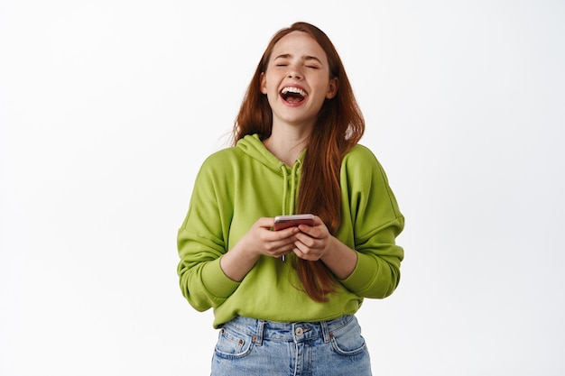 Happy redhead woman laughing, watching something funny on smartphone and having fun, enjoying video, standing in hoodie on white