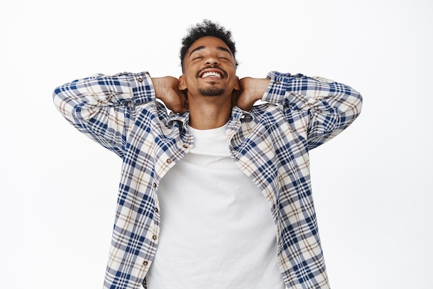 Happy and relaxed african american guy resting, close eyes and lay back, holding hands behind with joyful smile, relaxing on vacation, enjoy free time, white background.